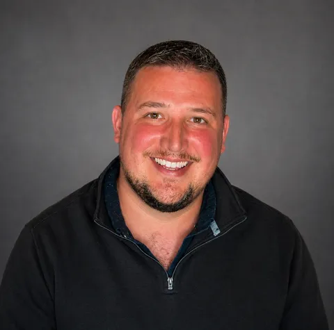 Drew Galbraith, smiling, dark zip pullover, neutral grey backdrop